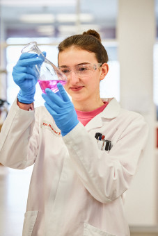 Eine Junge Frau, die eine Ausbildung zur Chemielaborantin macht, hält ein Gefäß mit pinker Flüssigkeit in der Hand.